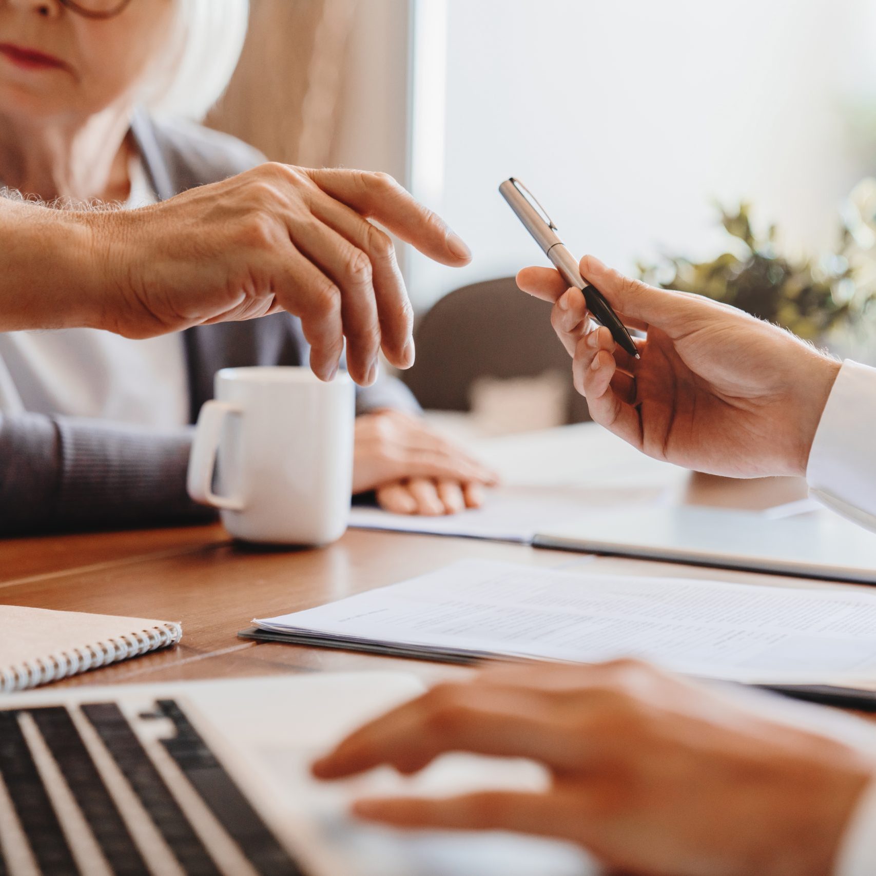 Käsi ojentaa kynää toiselle. Pöydällä on tietokone, kahvikuppi ja sopimusasiakirjoja.