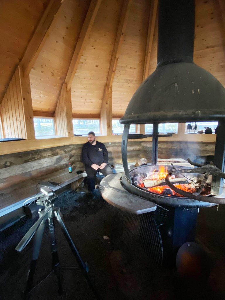 Lieksan omakotiyhdistyksen puheenjohtaja Aki Ikonen kodassa. Tulisijassa palaa nuotio, ja Ikosen eteen on viritetty kamera kuvaamaan.